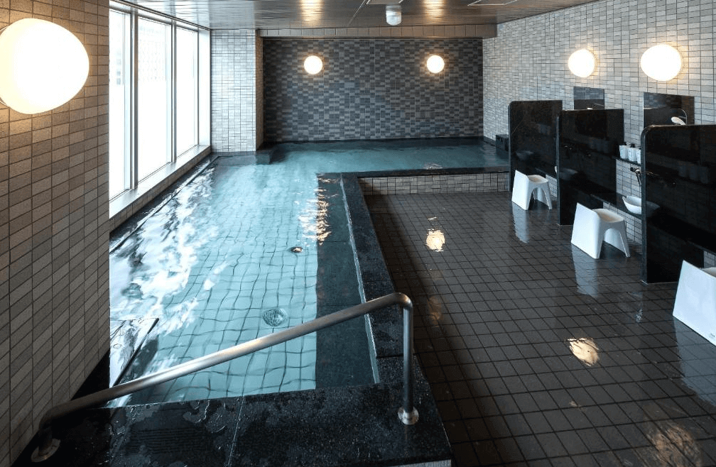 【旭川住宿】Y飯店-旭川站前 (Y's Hotel Asahikawa) 大浴場，設有寬敞室內溫泉池與洗浴區，採用灰白色磁磚裝潢，大片窗戶可引入自然光。