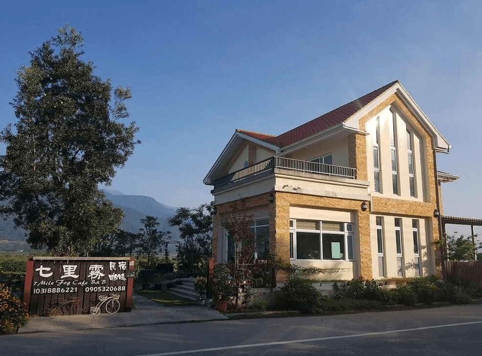 【赤柯山住宿】七里霧民宿外觀：歐風三角屋頂建築，白色與米黃磚牆搭配落地窗設計，門口設有招牌與綠意庭園，背後是山巒美景。