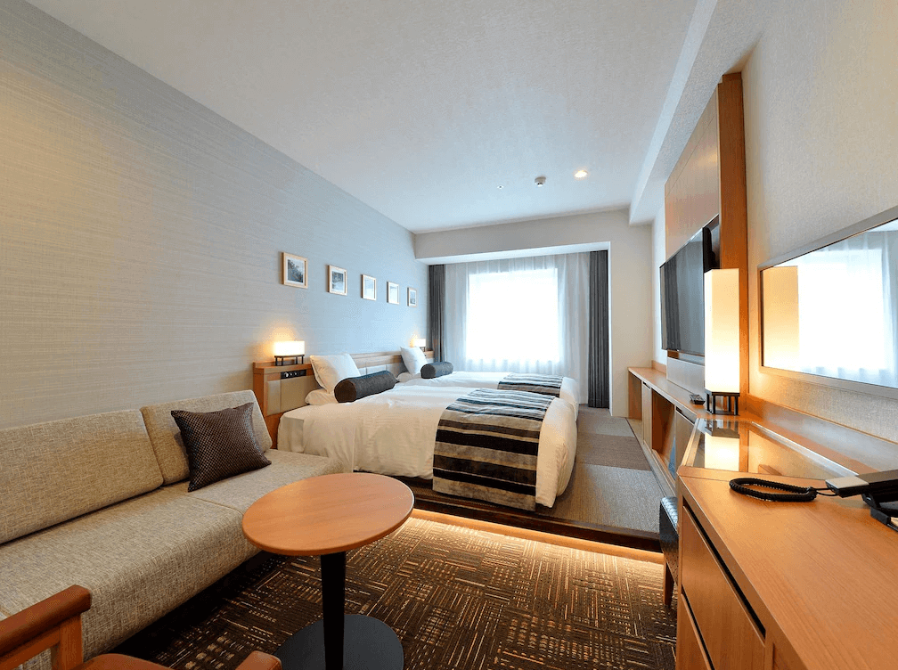 【函館住宿】函館國際酒店舒適雙床房，內部採用明亮木質設計，設有沙發與桌椅，窗外自然光灑落，營造溫馨氛圍。