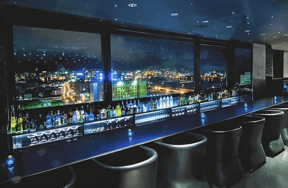 【函館住宿】函館普樂美雅凱賓總統酒店高樓層景觀酒吧，吧台前設有多種酒品，窗外能欣賞函館夜景與城市燈火。