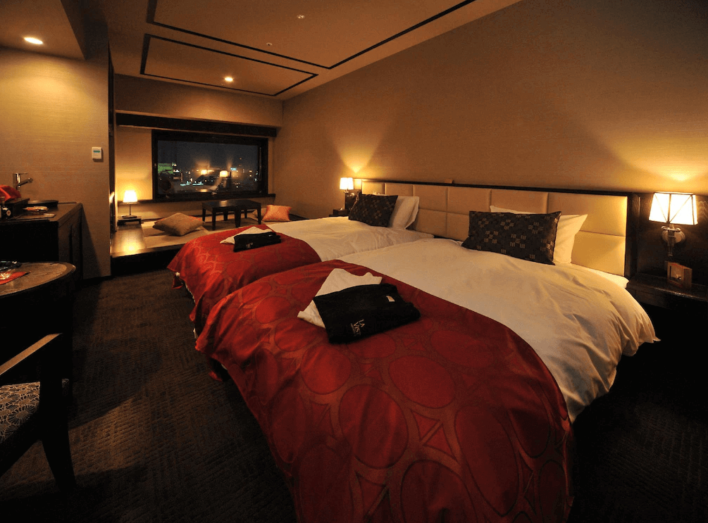 【函館住宿】函館灣 La Vista 飯店豪華雙床客房，紅色床罩點綴沉穩空間，窗邊設有觀景座位區，可欣賞夜晚的城市燈火。