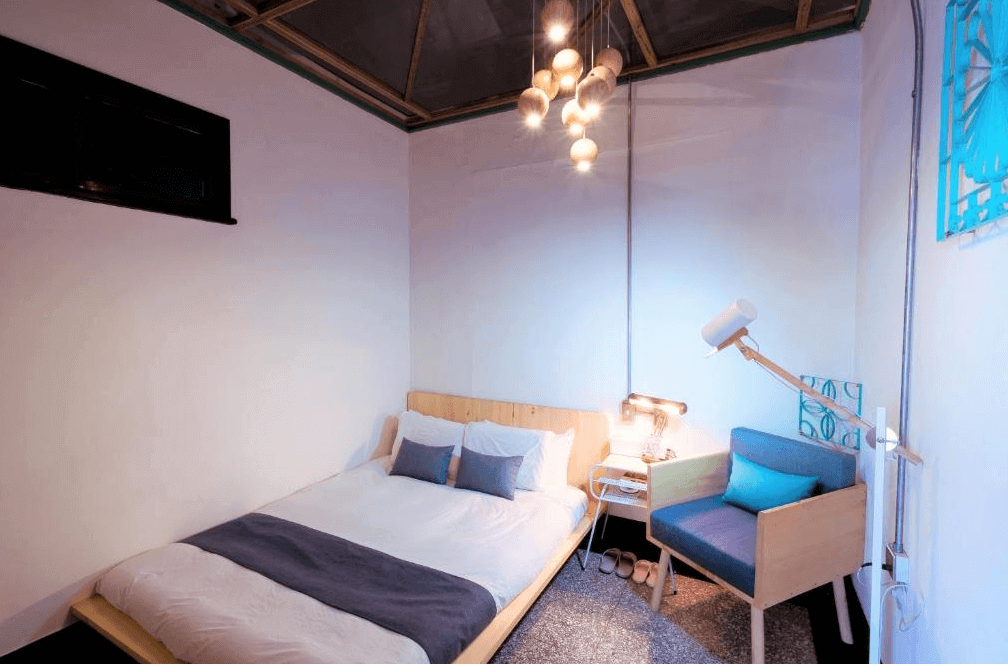 【高雄中央公園住宿】同．居 With Inn Hostel客房：溫馨雙人房，木質床架搭配白色床單與藍色抱枕，房內設有木質扶手椅與柔和吊燈，氛圍簡約溫暖。