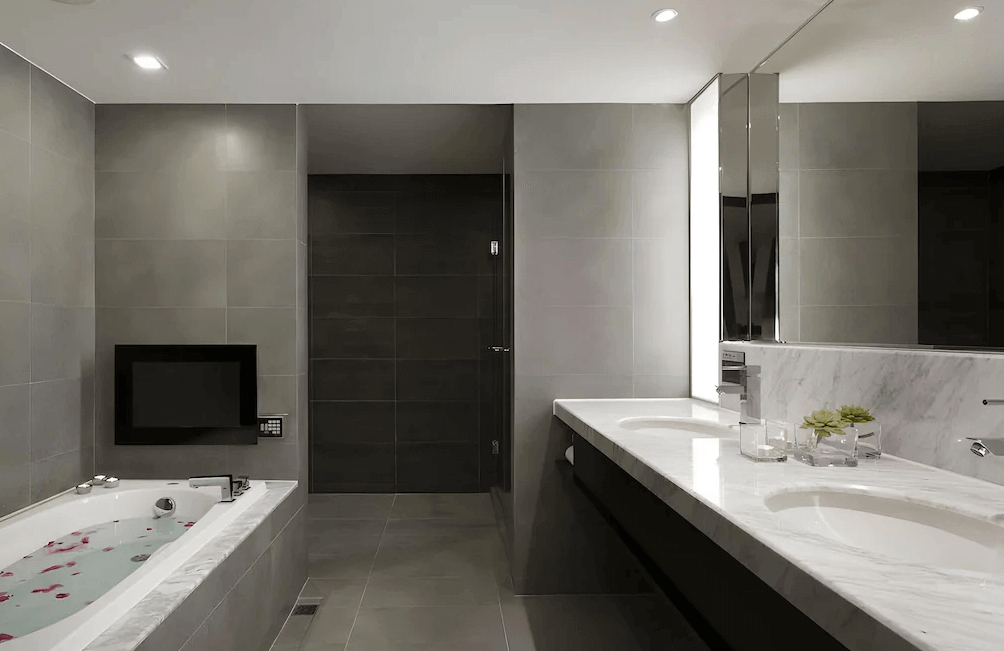 【高雄中央公園住宿】塔木德酒店高雄愛河館浴室：高質感灰色磁磚空間，設有浴缸與電視、雙人洗手台及大面鏡子，兼具時尚與實用。