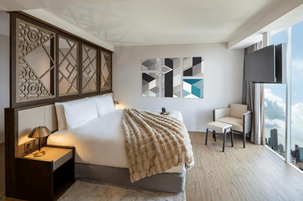 【高雄中央公園住宿】天成逸旅-舶寓高雄客房：溫馨大床房型，設有木質雕花床頭設計，大片落地窗可眺望城市天際線，室內搭配柔軟毛毯與現代風格裝飾畫。