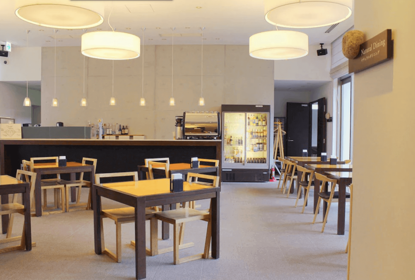 【富良野住宿】富良野Natulux飯店 (Furano Natulux Hotel) 餐廳用餐區，木質桌椅搭配簡約吊燈設計，空間明亮溫馨，後方設有吧檯與飲品區。