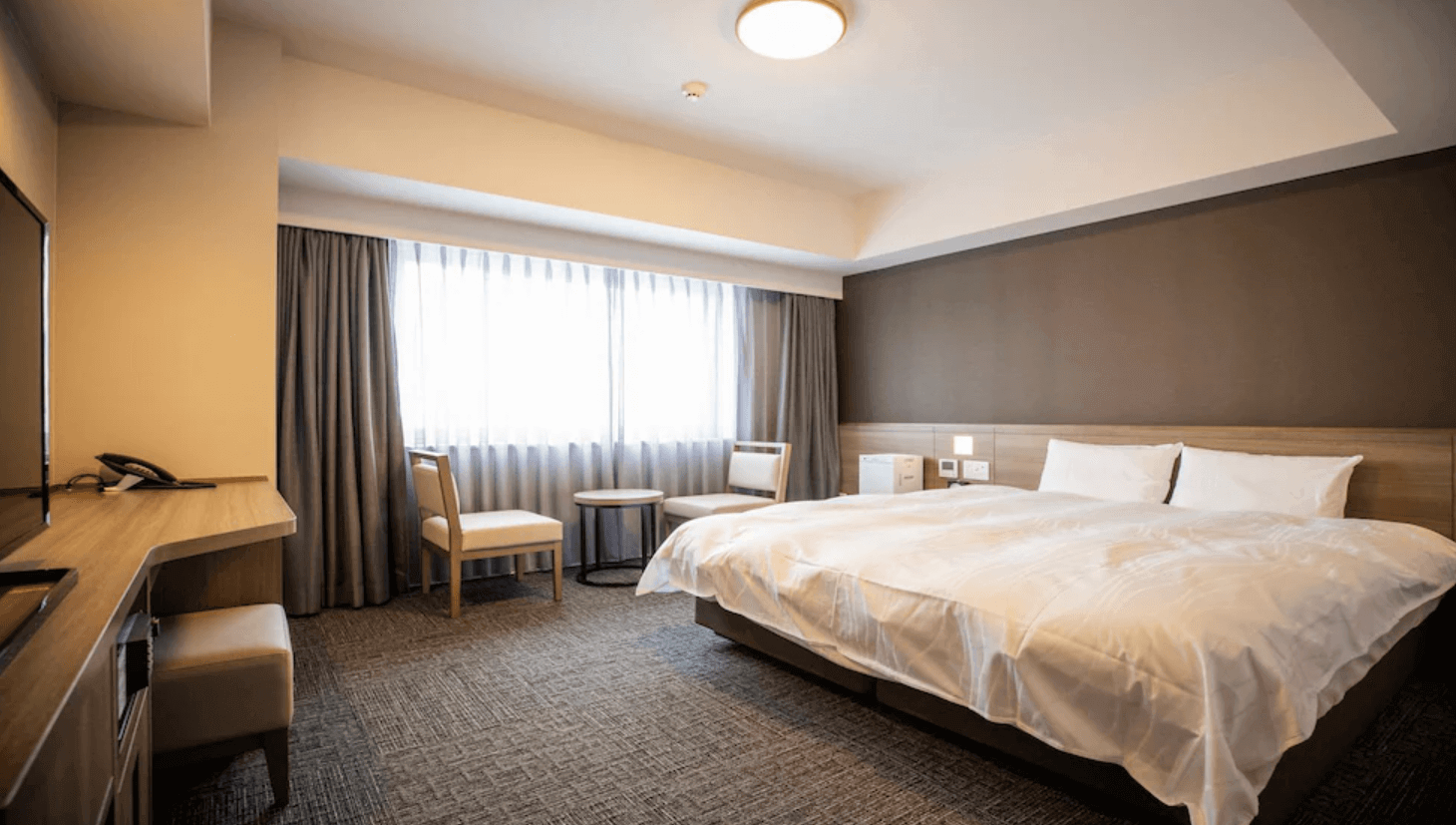 【富良野住宿】富良野拉維斯塔溫泉酒店 (La Vista Furano Hills Natural Hot Spring) 標準雙人房，房內設有一張大床、淺木色書桌、椅子與小圓桌，大片窗簾透出柔和自然光，整體空間簡約溫馨。