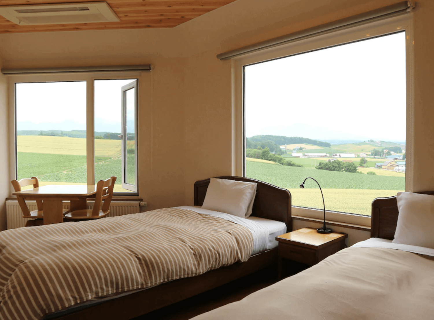 【富良野住宿】富良野風景民宿雙床房：房內擺放兩張單人床，大片落地窗外可欣賞遼闊的農田景色，並設有小桌椅供休憩。