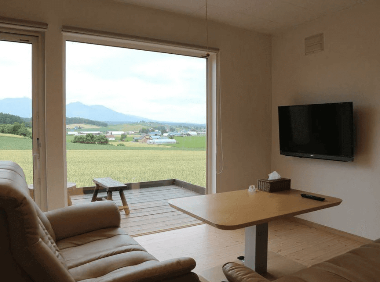 【富良野住宿】富良野風景民宿客廳：舒適沙發與木質餐桌，搭配大片窗戶可眺望田園風光，並有露台與電視設備。