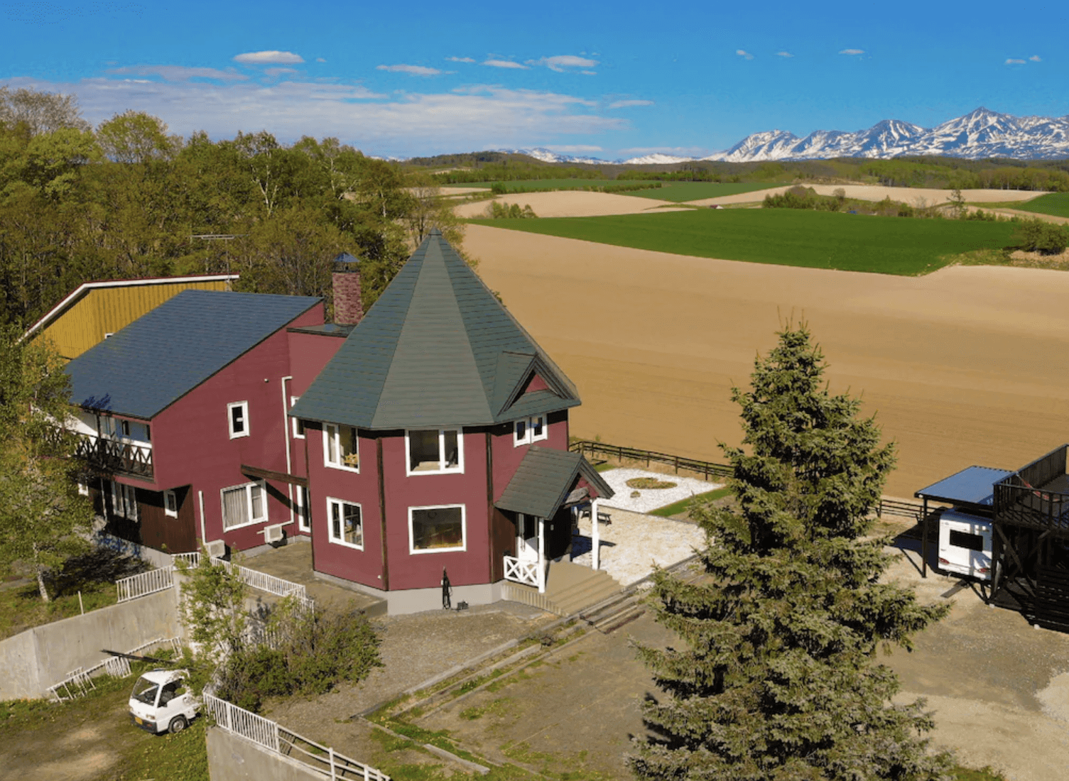 【富良野住宿】富良野風景民宿外觀：紅色建築搭配綠色屋頂，周圍環繞遼闊農田與遠方雪山，展現北海道鄉村特色。
