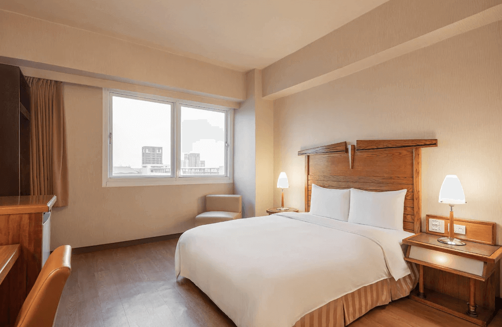 【鳳山住宿】康橋商旅五甲館雙人房溫馨簡約的雙人房，木質床頭與家具搭配白色床單，窗外可見高雄市景，整體氛圍明亮舒適。