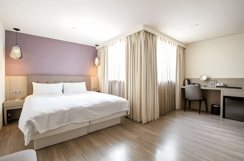 【三多商圈住宿】旅居文旅-亞灣館 標準雙人房：簡約設計風格，紫色牆面搭配大床，窗邊為書桌與茶水設施，空間寬敞乾淨。