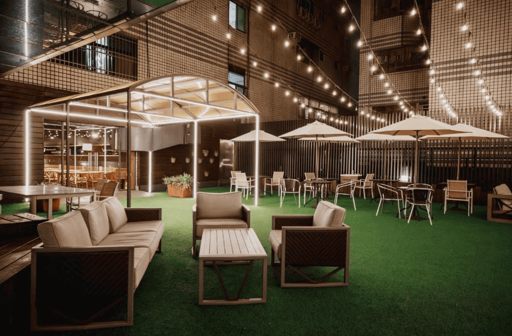 【三多商圈住宿】旅居文旅-亞灣館 戶外休憩區：夜間點綴串燈與陽傘座椅，綠色草坪搭配戶外沙發，營造愜意放鬆的氛圍。