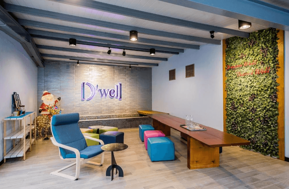 【三多商圈住宿】旅悅國際青年旅館交誼廳，牆面設有 D’well 標誌與綠植裝飾，搭配彩色坐墊與長桌，提供旅客交流與放鬆空間。