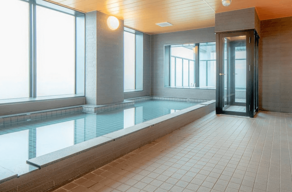 【旭川住宿】旭川HOTEL AMANEK (HOTEL AMANEK Asahikawa)公共浴場：室內大浴場以淺灰與木質色調打造，設有寬敞溫泉池與蒸氣室，大片窗戶讓光線灑落，氛圍放鬆舒適