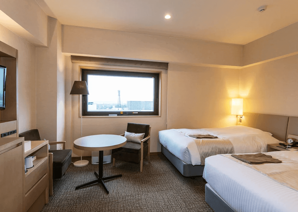 【旭川住宿】旭川JR飯店 (JR Inn Asahikawa) 雙床房，兩張單人床搭配柔和燈光，窗外可欣賞旭川市景，並設有小型桌椅，適合雙人入住。