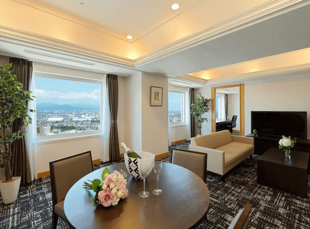 【札幌住宿】札幌Emisia飯店 豪華套房，寬敞客廳配有餐桌與沙發區，大窗外可遠眺城市與群山美景。