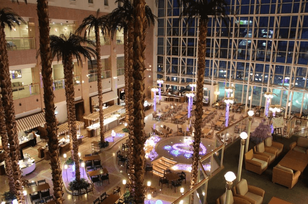 【札幌住宿】札幌朵蕾絲SPA度假飯店 中庭餐飲區，高挑玻璃帷幕設計，搭配棕櫚樹與燈光裝飾，展現度假氛圍。