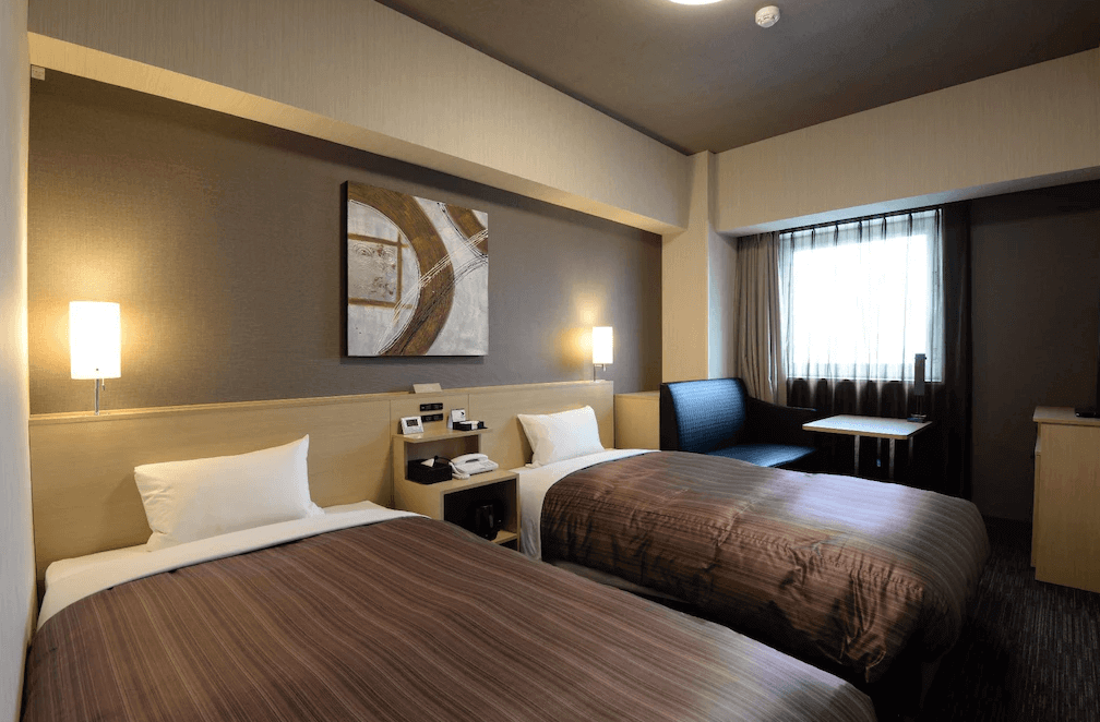 【旭川住宿】露櫻GRAND酒店旭川站前店 (Route Inn Grand Asahikawa Ekimae) 雙床房，現代簡約設計，兩張單人床搭配暖色燈光與牆面藝術畫，營造舒適氛圍。