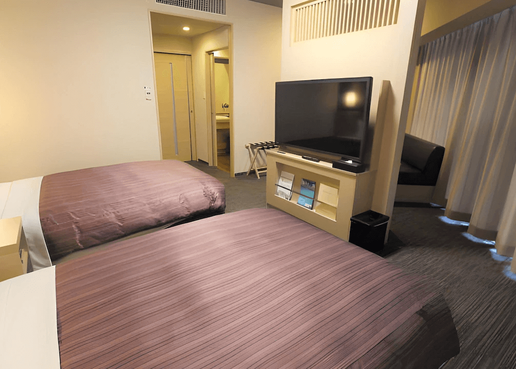 【旭川住宿】露櫻GRAND酒店旭川站前店 (Route Inn Grand Asahikawa Ekimae) 客房內部，設有大螢幕電視、兩張單人床與沙發休憩空間，溫馨舒適。