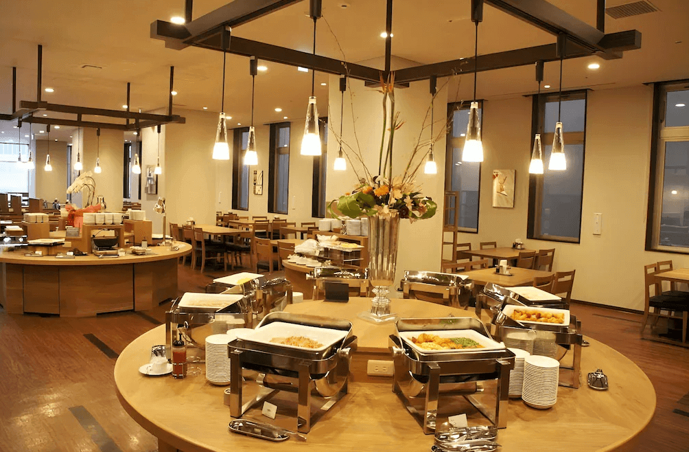 【旭川住宿】露櫻GRAND酒店旭川站前店 (Route Inn Grand Asahikawa Ekimae) 餐廳自助早餐區，木質桌椅搭配明亮燈光，餐檯擺放多樣料理與新鮮食材，提供豐盛早餐。
