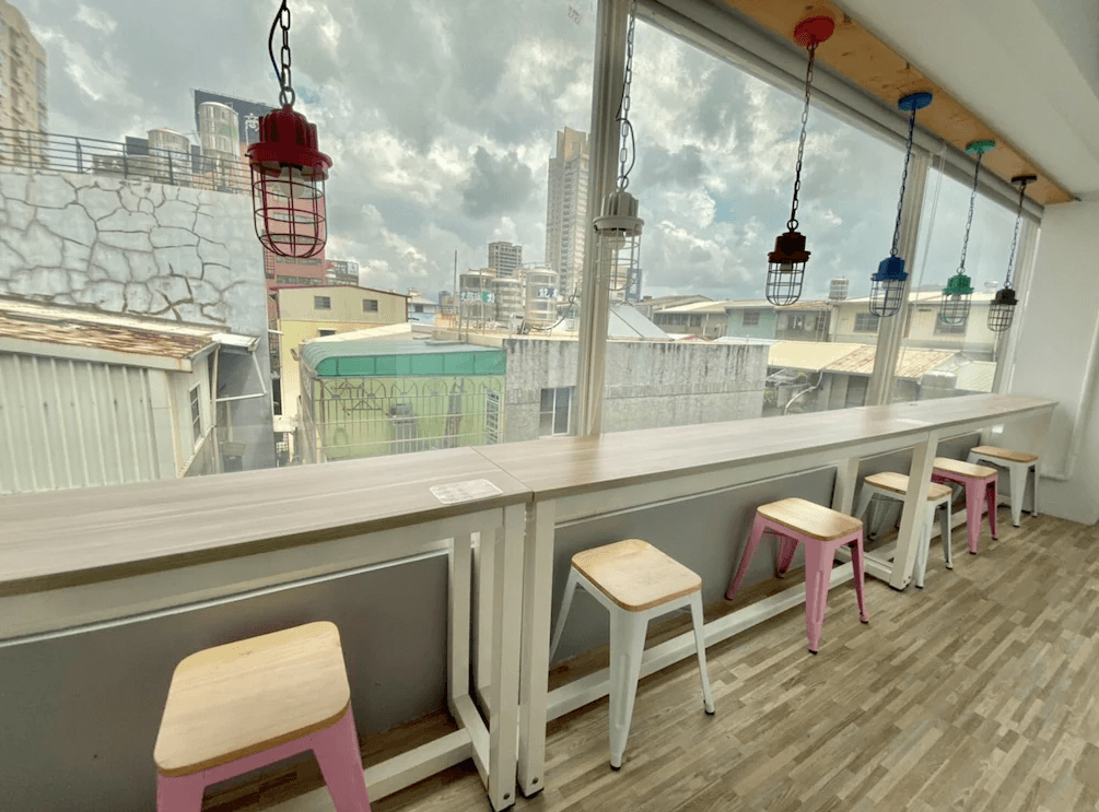 【三多商圈住宿】飛行家青年旅館 FLYINN HOSTEL 公共空間，面向落地窗的吧檯式座位，搭配多彩吊燈與高腳椅，可欣賞高雄市景。