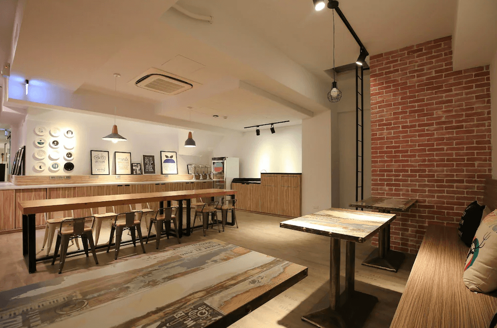 【三多商圈住宿】飛行家青年旅館 FLYINN HOSTEL 交誼廳，磚牆與木質設計營造工業風氛圍，設有長桌與沙發區，提供旅客交流休憩。