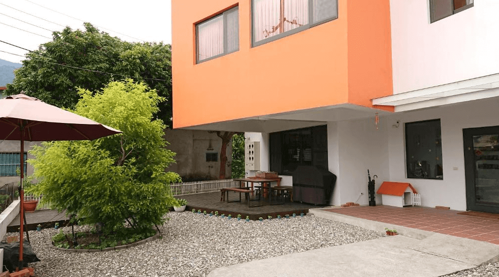 【赤柯山住宿】高寮貳陸之貳民宿外觀：建築外牆以白色與橘色相間設計，庭院內鋪設碎石與木棧板，設有遮陽傘與戶外座椅，環境清幽舒適。