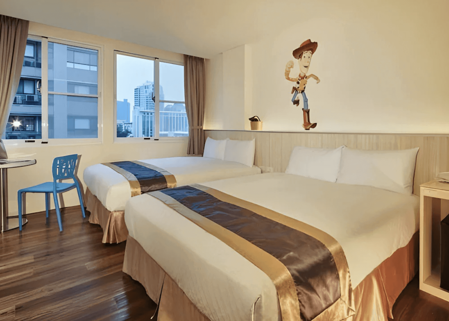 【前金住宿】高雄都會商旅 M-Hotel 客房，內有兩張雙人床，牆面上有卡通牛仔裝飾，大片窗戶可眺望市景，空間溫馨明亮。