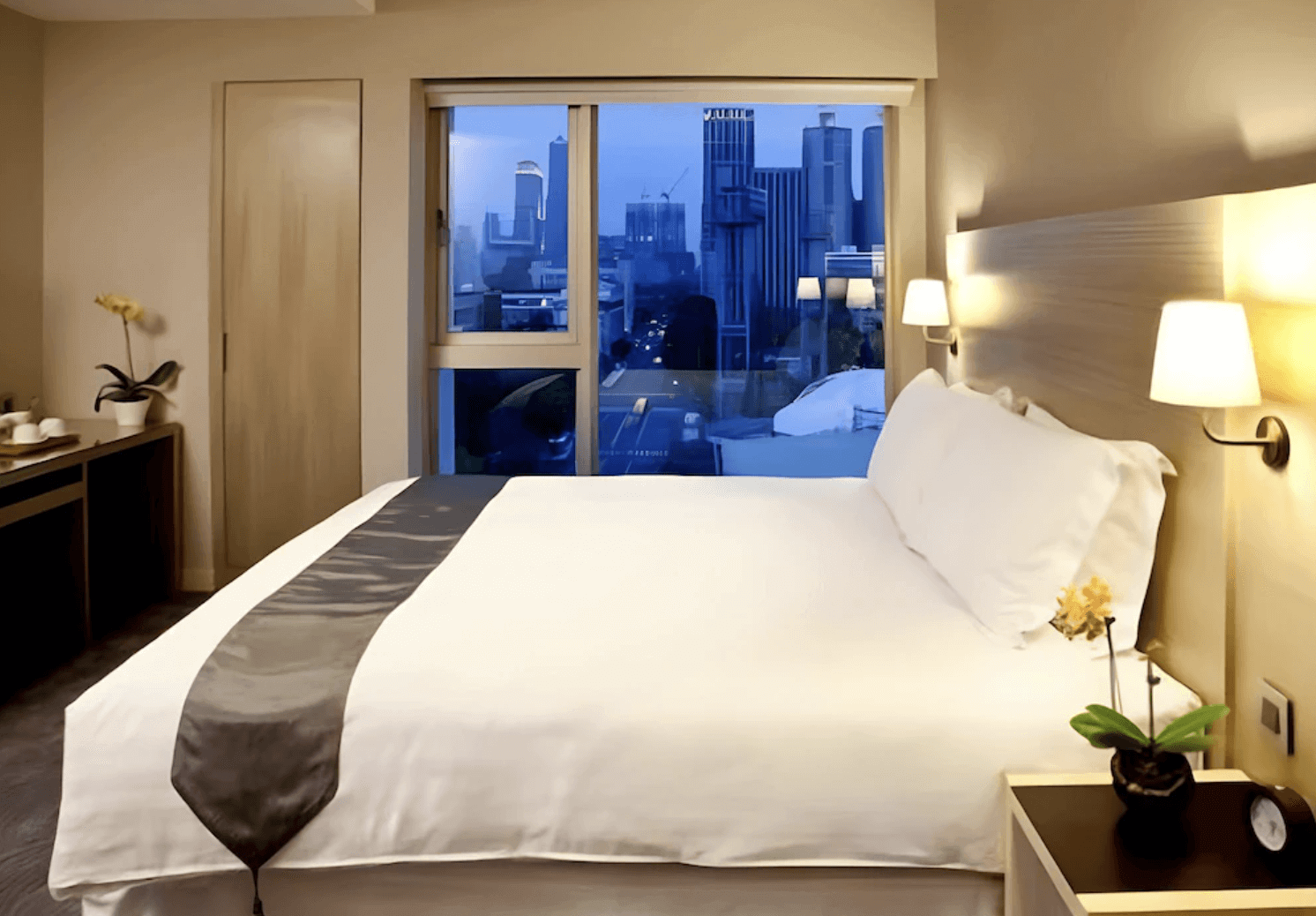 【前金住宿】麗馨精品商旅七賢館 — 夜晚客房氛圍溫馨，落地窗外可見高雄城市夜景，床頭燈光柔和。