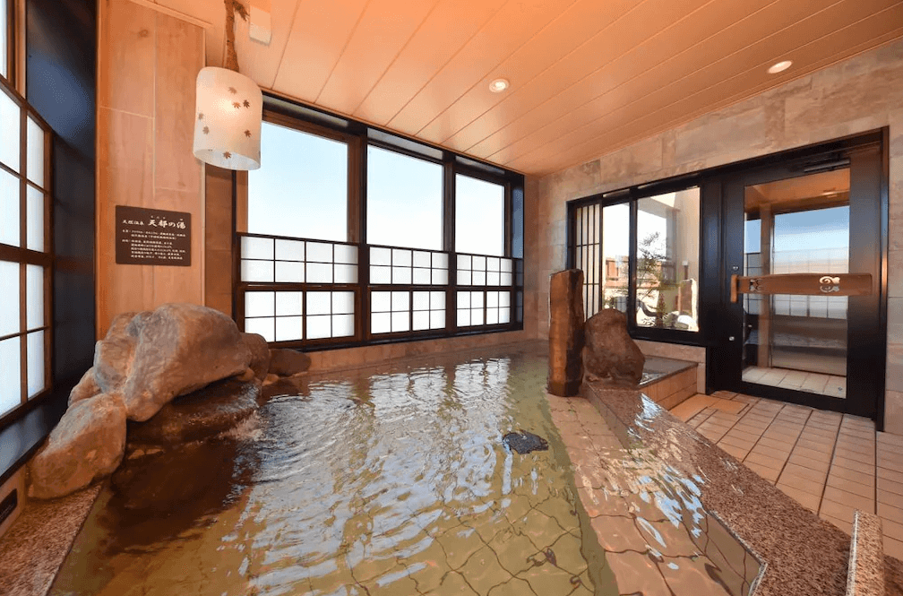 【網走住宿】「Dormy Inn 網走天然溫泉飯店」的室內天然溫泉浴場，採大面窗景設計，浴池以岩石與木質元素打造，可邊泡湯邊欣賞窗外景色，旁設有桑拿房入口。