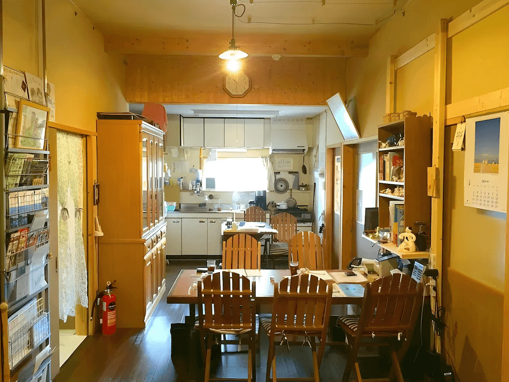 【網走住宿】Imodango村民宿」的公共餐廳與廚房空間，採木質家具與柔和燈光，牆上擺滿旅遊資訊與書籍，整體呈現溫馨舒適的家庭式氛圍。