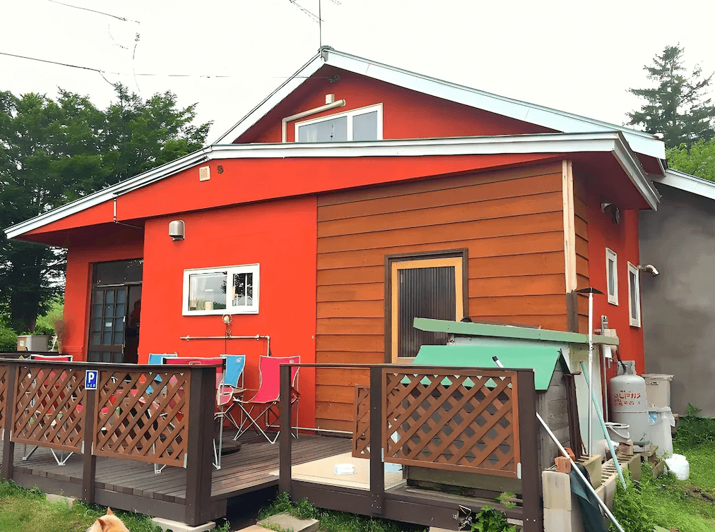 【網走住宿】Imodango村民宿」的外觀建築，鮮豔橘紅色外牆搭配木質圍欄與露台座椅，周圍綠意環繞，展現溫馨可愛的鄉村風格住宿環境。