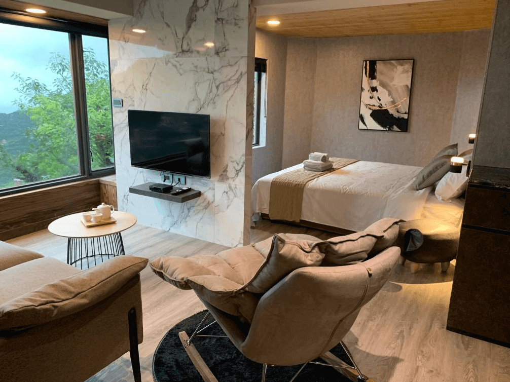 【瑞芳住宿】九份山城逸境｜景觀套房一景，採光明亮的落地窗可遠眺山巒綠意，房內設有沙發區、電視與現代風設計牆面，營造溫潤質感的度假氛圍。