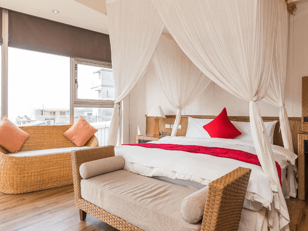 【瑞芳住宿】九重町客棧｜浪漫四柱床雙人房，設有白色紗幔與紅色抱枕點綴，藤編沙發與木質地板營造溫暖自然氛圍，窗邊採光明亮。