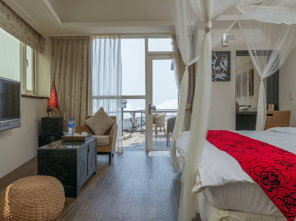 【瑞芳住宿】九重町客棧｜海景套房設有紗幔大床與獨立陽台，房內以自然藤編家具與米色窗簾搭配，可從陽台遠眺九份山海景色。