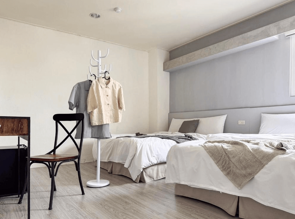 【台南寵物友善住宿】信然文旅・首學的雙床房，以灰白色牆面與木質地板呈現簡約設計風格，房內設有衣架與書桌椅，空間整潔明亮，適合家庭或旅人入住。