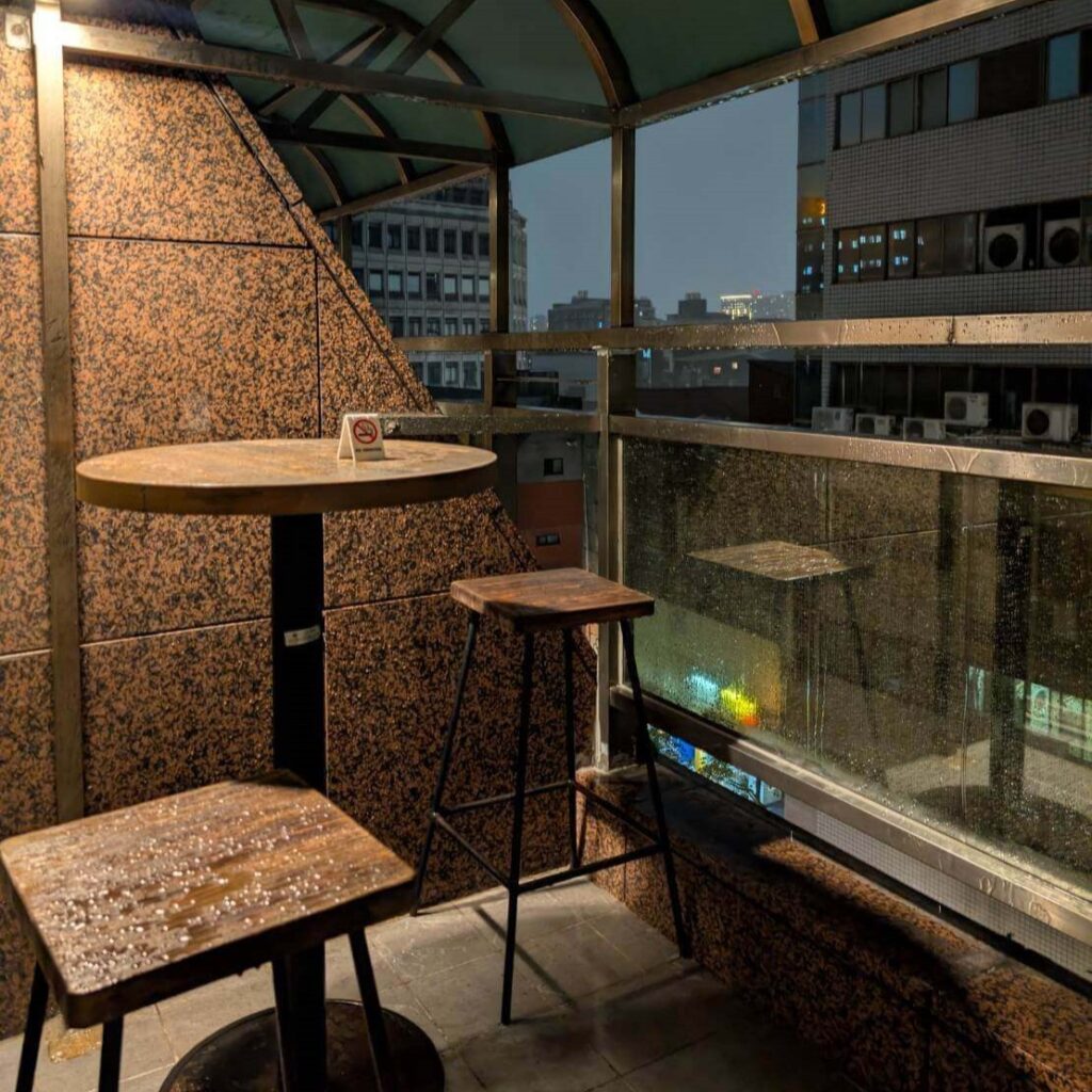 【台北背包客棧推薦】北門臥客青年旅館陽台夜景區，設有高腳桌與椅的露天陽台，可俯瞰市區夜景，雨後地面微濕，氣氛靜謐放鬆。