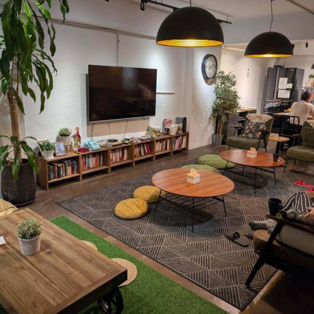 【台北背包客棧推薦】北門臥客青年旅館公共交誼廳，溫馨開放式交誼空間，配有電視、書架、綠植、圓桌與坐墊，整體氛圍溫暖且具文青風格。