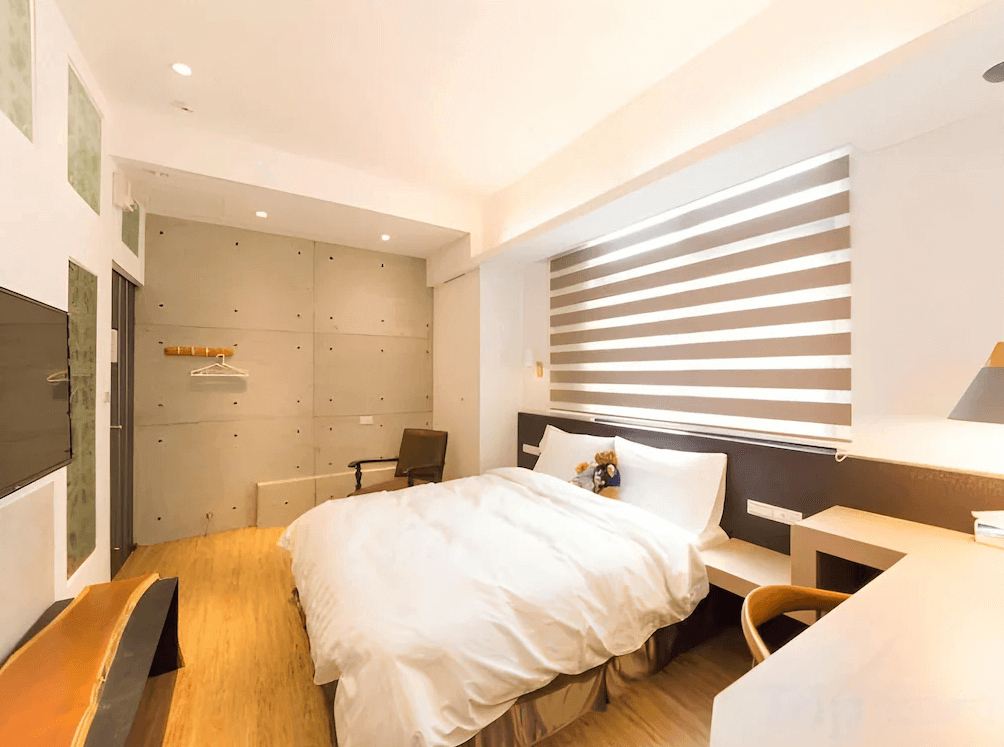 【台南寵物友善住宿】台南民宿湧福驛站的雙人房，房內以明亮木質色調為主，簡約設計結合工業風混凝土牆面，搭配整齊舒適的大床與百葉窗簾，營造溫馨又現代的住宿氛圍。