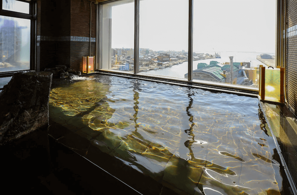 【釧路住宿】多米高級酒店－釧路天然溫泉頂樓展望浴池，可眺望釧路港灣與城市景觀，陽光灑落在溫泉水面，景致療癒放鬆。