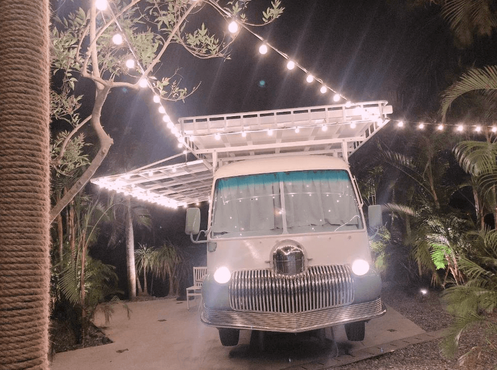 【頭城住宿】宜蘭工匠家小木屋民宿 露營車夜景，車體外掛滿串燈飾，周圍被綠意與棕櫚樹環繞，營造浪漫露營氛圍。