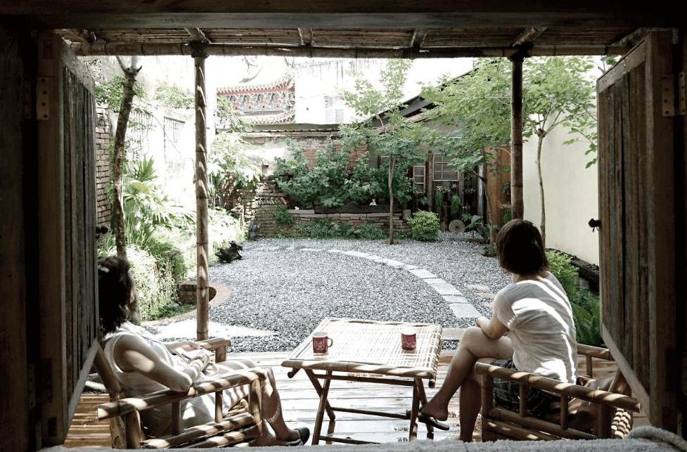 【台南寵物友善住宿】巷弄 x 台南民宿庭院景觀，住客坐在竹椅上享受早晨陽光，庭院以碎石小徑與綠意植栽打造，展現老宅靜謐氛圍。