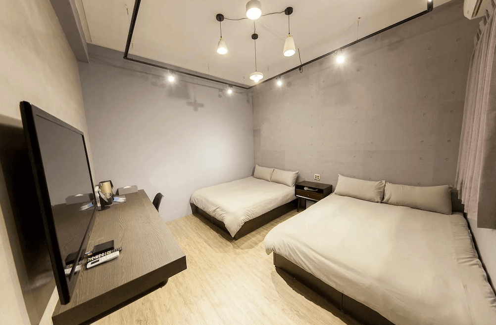 【台南寵物友善住宿】怠宿 SLOW 四人房，簡約水泥灰牆與木質地板設計，設有兩張雙人床與吊燈照明，整體風格極簡又具工業感。