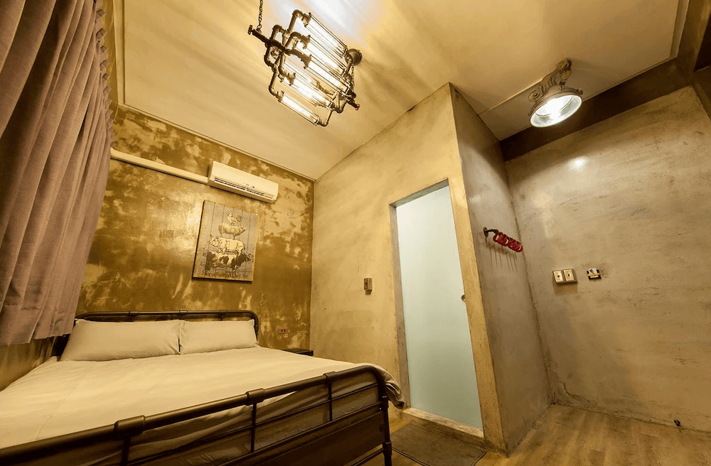 【台南寵物友善住宿】怠宿 SLOW 雙人房，以金屬床架與復古吊燈展現工業風格，牆面以仿舊金色塗層搭配掛畫，呈現沉穩質感氛圍。
