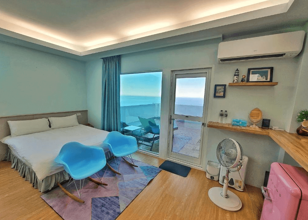 【瑞芳住宿】應許美地民宿｜海景雙人房內部設計，木質地板、藍色搖椅與落地窗外可見無敵海景，房內配置冷氣與粉色小冰箱，整體風格清新舒適。