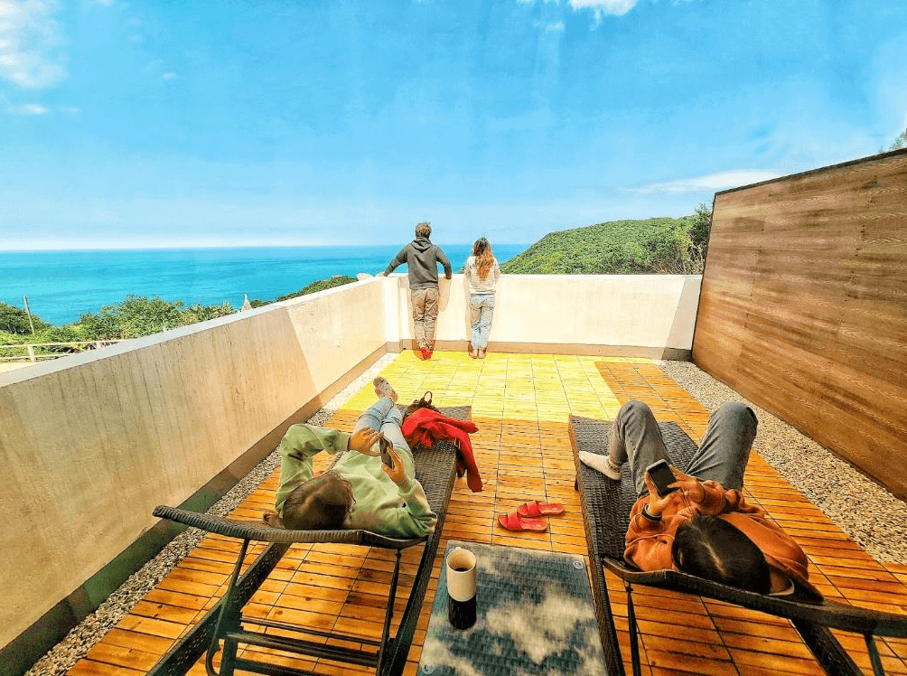 【瑞芳住宿】應許美地民宿｜旅客在海景露台上放鬆曬太陽、遠眺湛藍海岸，設有躺椅與木質地板，展現愜意度假氛圍。