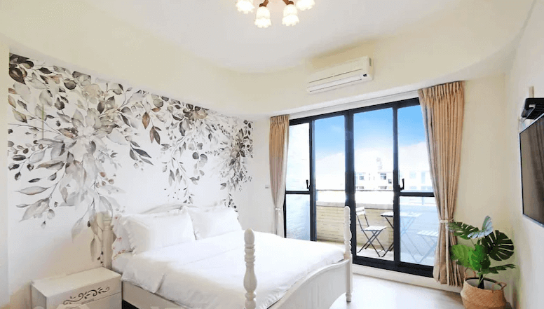 【枋寮住宿】「枋寮帝寶16民宿 雙人房，牆面以花卉壁畫裝飾，搭配白色床架與大面落地窗，窗外設有小陽台與座椅，可遠眺藍天與屋頂景色。」