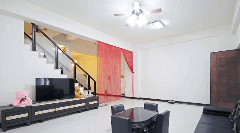 【枋寮住宿】「枋寮帝寶16民宿 客廳空間寬敞明亮，設有電視、沙發與餐桌，紅色布簾與熊玩偶增添溫馨感，適合家庭或團體入住。」