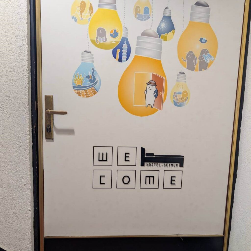 【台北背包客棧推薦】北門臥客青年旅館入口門設計，旅館房門上以多個黃色與藍色燈泡插畫裝飾，內含可愛人物圖樣，下方印有「WELCOME」與「HOSTEL BEIMEN」字樣，充滿創意與活力。