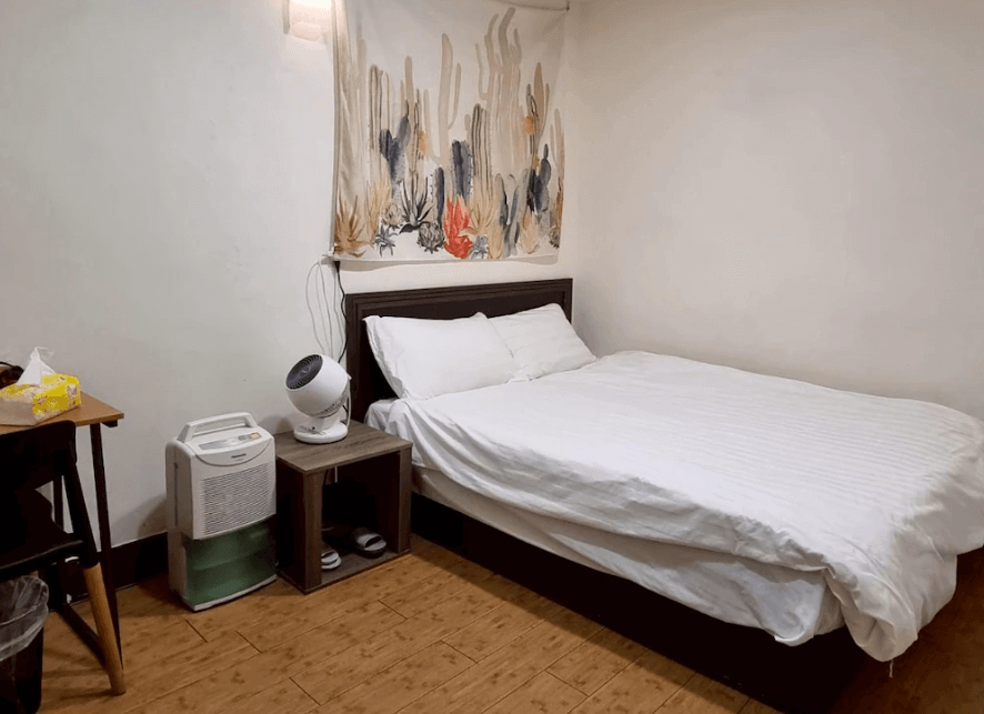 【瑞芳住宿】清芳民宿雙人房，床鋪柔軟潔白，牆上掛有抽象風格掛布裝飾，旁邊設有除濕機與電風扇，整體空間乾淨溫馨，適合長住旅客。